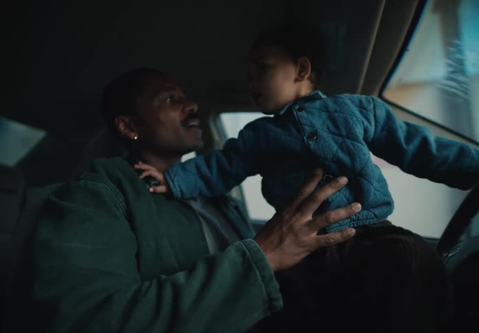 A joyful toddler sits on his father's lap, playfully steering the car as the dad watches with a loving smile