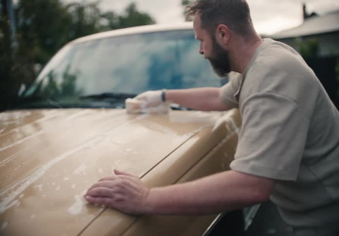 A bearded man meticulously scrubs the hood of his vintage tan pickup truck with a soapy sponge in slow motion