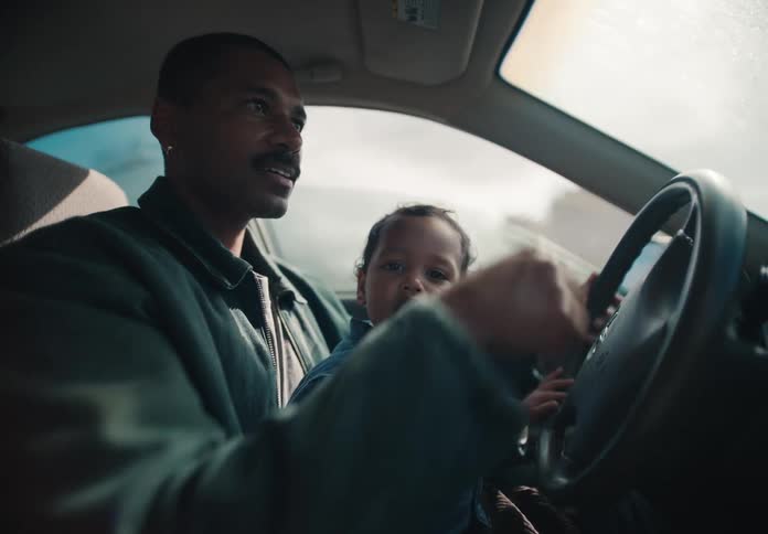 An African American father drives his car with his toddler on his lap, talking to them as they look out the window together
