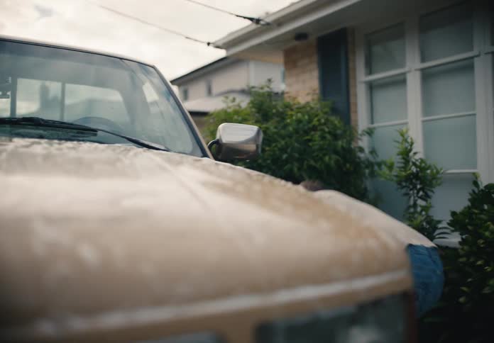 A father and son playfully wash a vintage truck in their driveway, engaging in a fun water fight with sponges and a hose