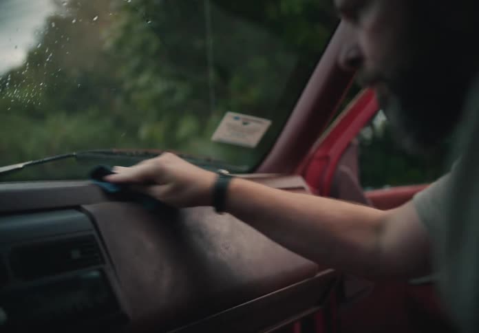 A bearded man carefully wipes dust from the dashboard of his vintage red truck parked outdoors