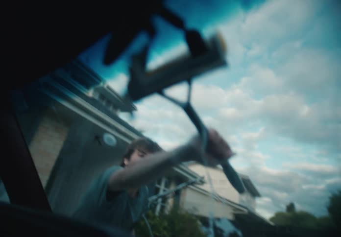 From inside a car, a young man in a t-shirt methodically cleans the soapy windshield with a squeegee in slow motion