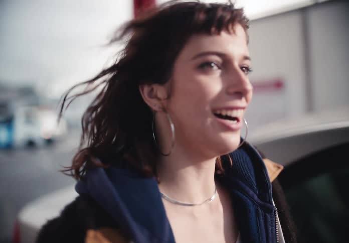 A cool young woman with windblown hair smiles as she walks confidently through an urban parking lot in slow motion