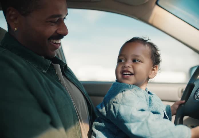 Happy toddler sits on his smiling father's lap in the driver's seat, pretending to drive the car on a sunny day