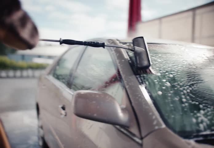 A young man scrubs the soapy windshield of his car with a long brush at a self-service car wash on a sunny day
