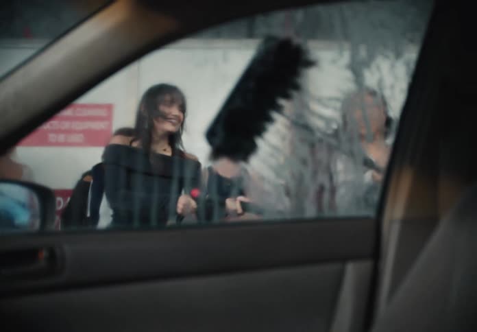 A group of friends playfully wash a car window with a pressure washer and brush, filmed in slow motion from inside the vehicle