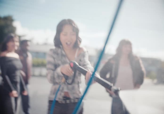A smiling young woman playfully aims a pressure washer while her friends watch at a sunny outdoor car wash in slow motion