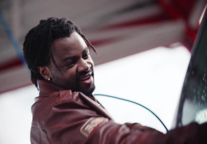 A smiling man in a leather jacket power washes the windshield of his car in slow motion at a self-service car wash