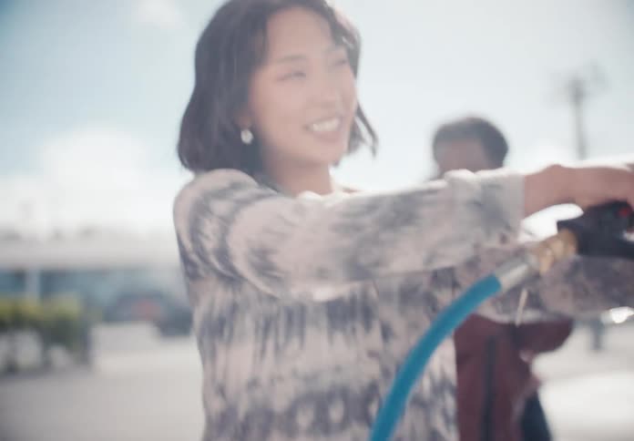 Slow motion close-up of a smiling Asian woman playfully using a pressure washer at a car wash on a bright, sunny day