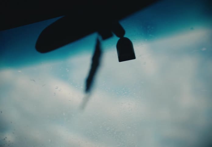 A person washes the exterior of a car windshield, as seen from the inside looking out at the sky