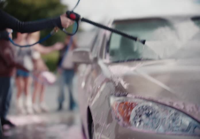 A stylish young woman in a black top uses a pressure washer to rinse pink soap off a car at a self-service bay in slow motion