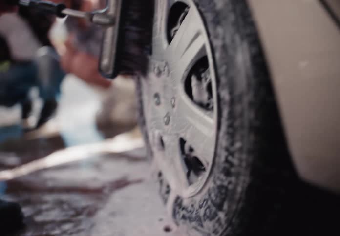 A man scrubs a soapy car tire and rim with a long brush in slow motion at a car wash