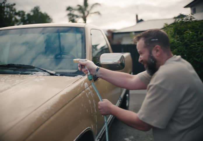 A happy bearded man scrubs his classic tan pickup truck with a soapy sponge before rinsing it off with a hose in his driveway, shot in slow motion