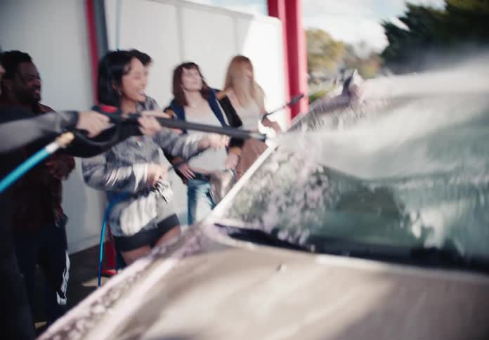 Diverse group of friends joyfully laugh and play while washing a car at a self-service car wash in slow motion