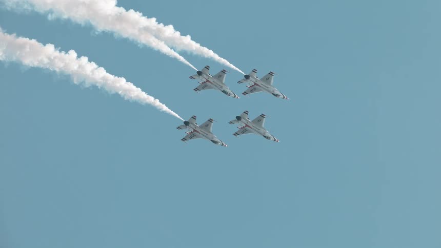 Four fighter jets fly in perfect formation across a clear blue sky, leaving long white smoke trails in a dramatic slow-motion shot