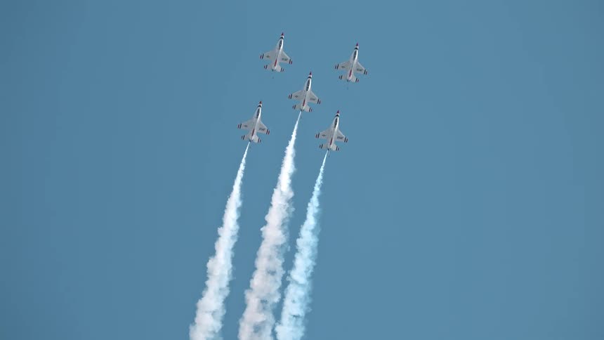 A squadron of five aerobatic jets performs a synchronized climb in slow motion, trailing white smoke across a brilliant blue sky