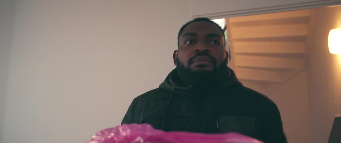A bearded man walks with determination while carrying a box and a pink bag through a hallway in slow motion