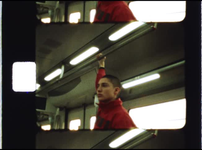 A young person with a shaved head in a red sweatshirt holds onto an overhead bar while riding on a train, captured on vintage film