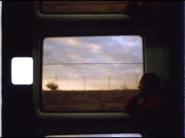 Vintage super 8mm footage shows a young boy in a red shirt looking out of a train window as the countryside speeds by at sunset