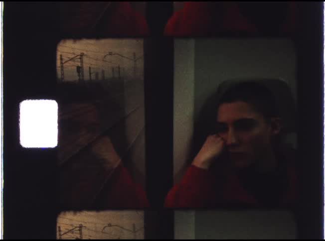 A pensive person in a red sweater sits on a train as their reflection appears in the window next to passing tracks in this vintage film footage