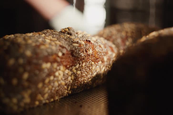 Close-up of a baker sliding a tray of freshly baked artisan bread with seeds in a warm bakery