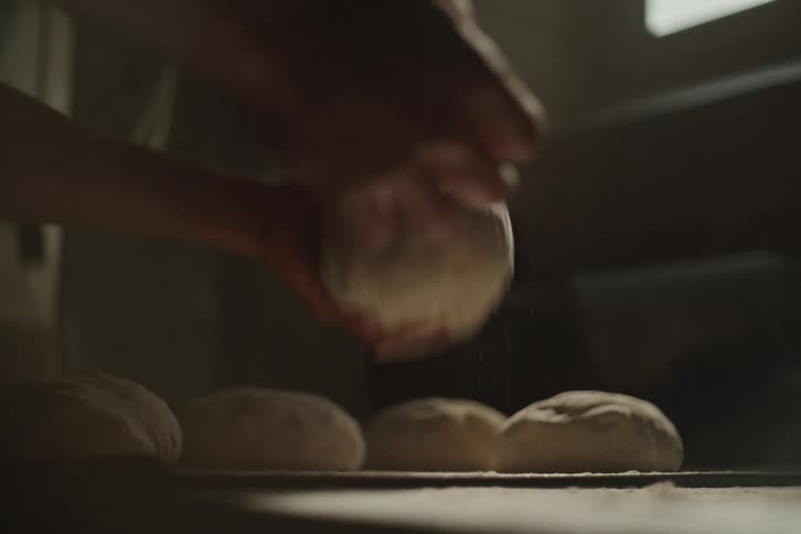 A baker's hands gently dust a ball of dough with flour in slow motion in a dimly lit kitchen