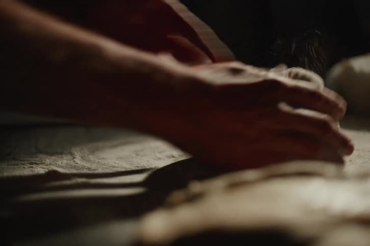 A baker's hands knead and shape dough on a floured surface in a dimly lit, rustic kitchen