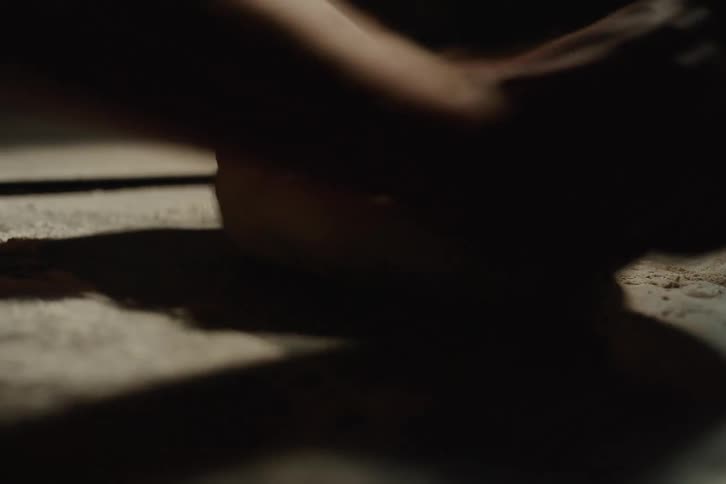 A baker's hands knead dough on a floured surface in a dimly lit, rustic kitchen