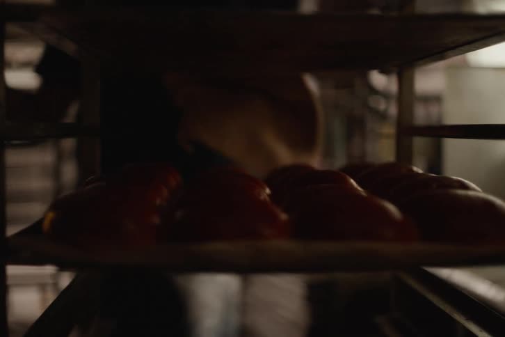 A baker slides trays of dough into a large industrial oven, panning across racks of freshly baked golden-brown buns cooling in a warm, atmospheric bakery