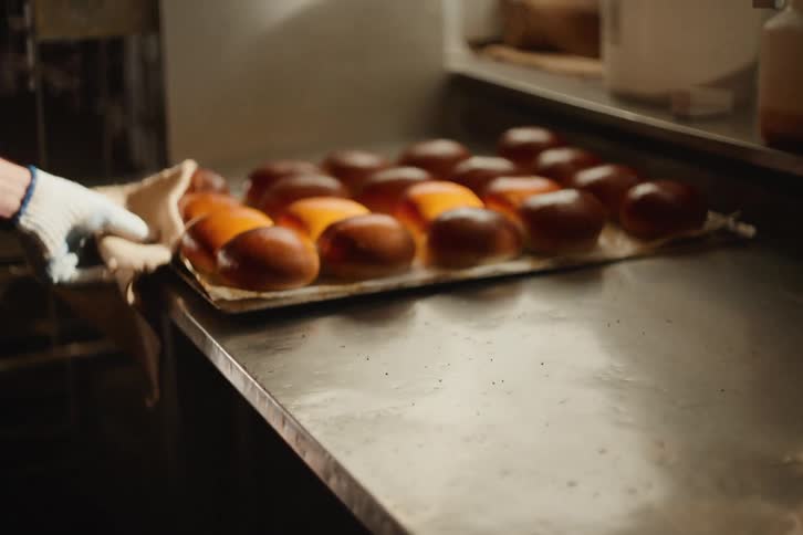 A baker in slow motion places a tray of freshly baked, warm brioche buns onto a stainless steel kitchen counter