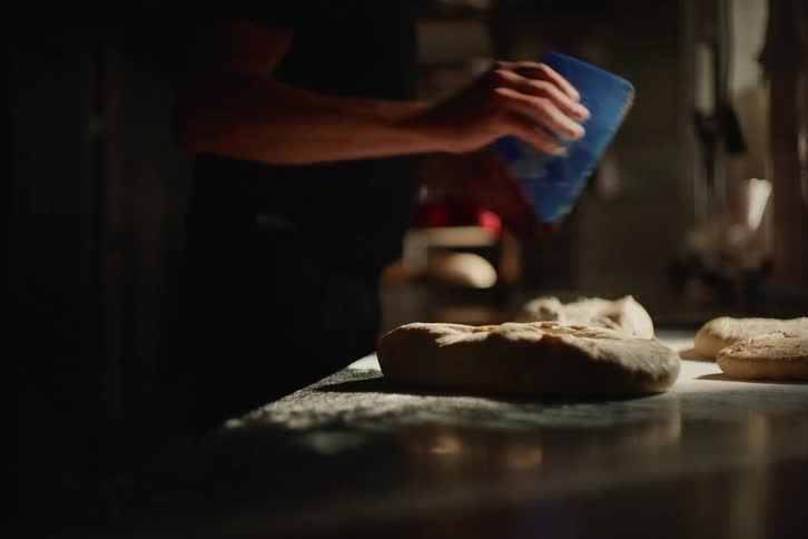 An artisan baker expertly shapes a loaf of bread dough on a floured countertop in a dimly lit kitchen in slow motion
