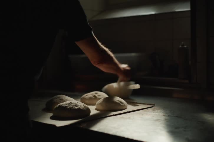 A baker dusts fresh dough balls with flour in a dimly lit kitchen before putting them in the oven in slow motion