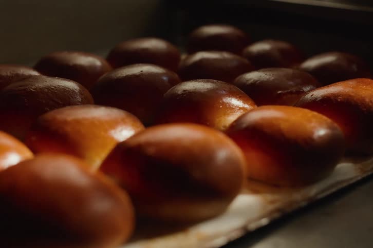 A close-up slow dolly shot reveals rows of freshly baked, golden-brown brioche buns on a baking tray