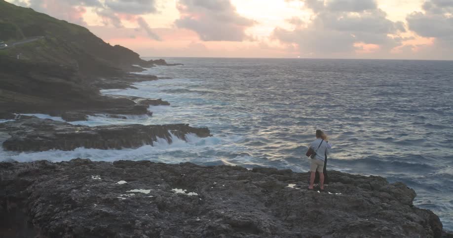 An aerial reveal shows a romantic couple embracing on a dramatic, rocky shoreline as powerful waves crash below them at sunrise