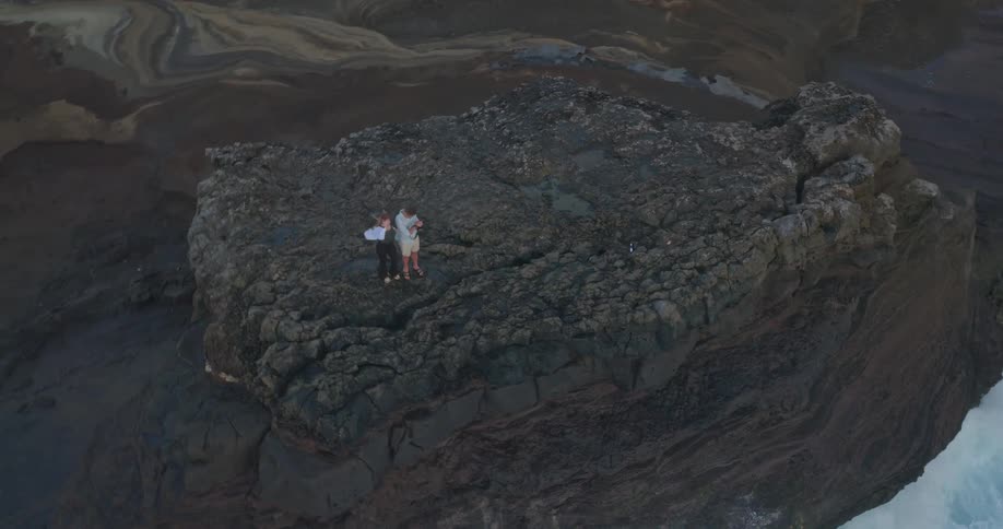 An aerial view shows a romantic couple embracing on a rugged coastal cliff as the drone slowly ascends, revealing the powerful ocean waves crashing below