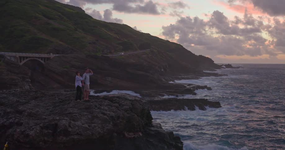 A loving couple takes a selfie and admires the beautiful sunrise from a dramatic, rocky coastline