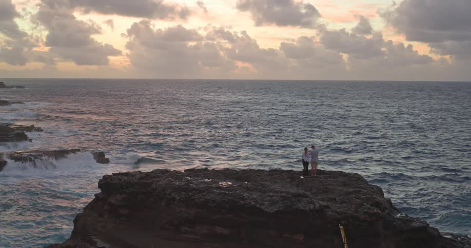 A drone pushes in on a couple watching the sunrise from a rocky ocean cliff, then pans to reveal the vast, rugged coastline