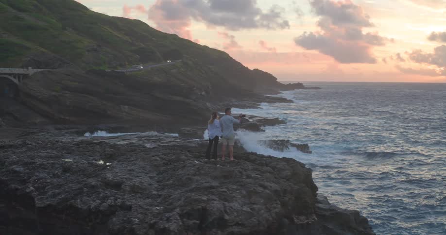 A slow zoom out reveals a couple holding hands and taking photos on a rugged cliffside as waves crash below during a beautiful sunset