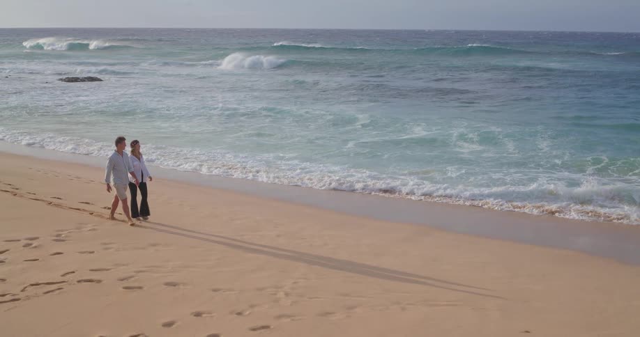 A serene slow-motion wide shot of a couple holding hands and walking along a beautiful sandy beach next to the ocean