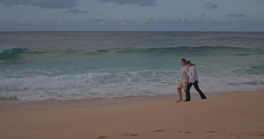 A romantic couple holds hands while walking on a beautiful sandy beach next to the ocean waves in slow motion