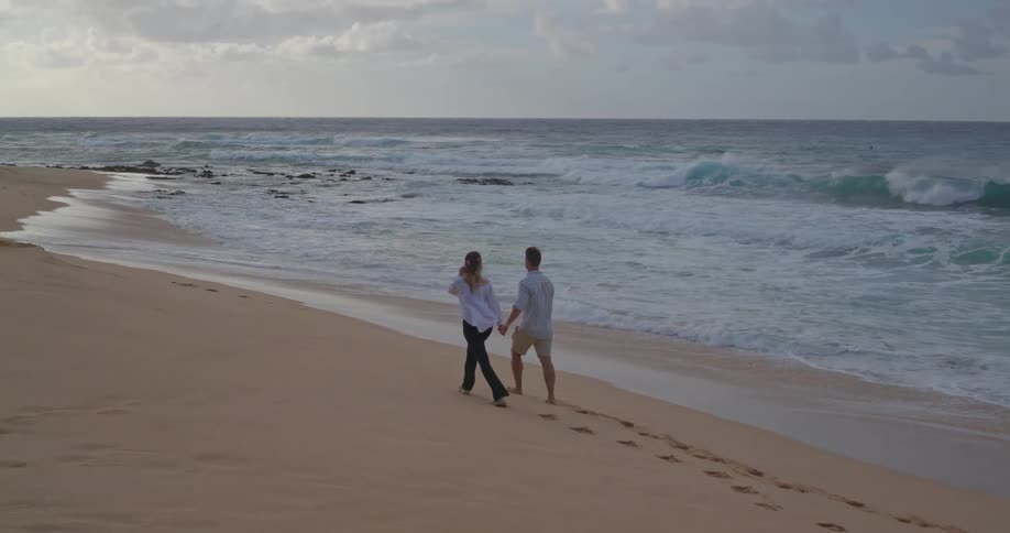 A couple walks hand-in-hand in slow motion along a deserted sandy beach, leaving footprints behind them as ocean waves roll ashore