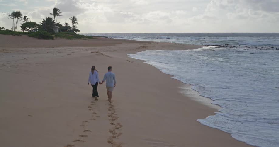 A romantic couple holds hands while walking barefoot along a beautiful sandy beach as the ocean waves crash on the shore