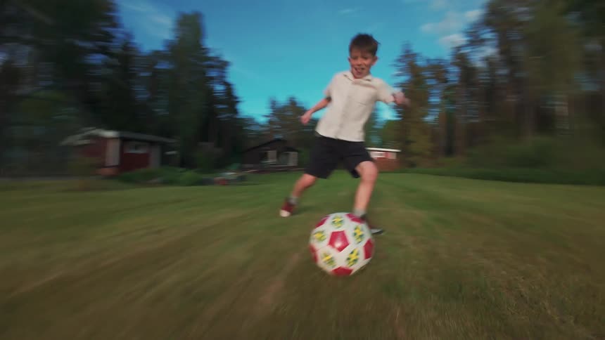 Dynamic slow-motion FPV footage follows a happy boy dribbling a soccer ball in a grassy backyard before kicking it towards the camera
