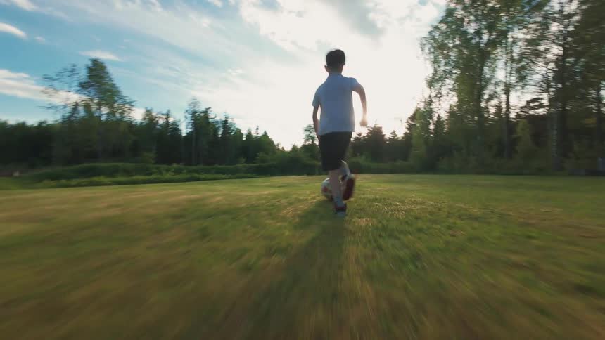 Dynamic low-angle follow shot of a boy running with a soccer ball across a green lawn during a beautiful sunset