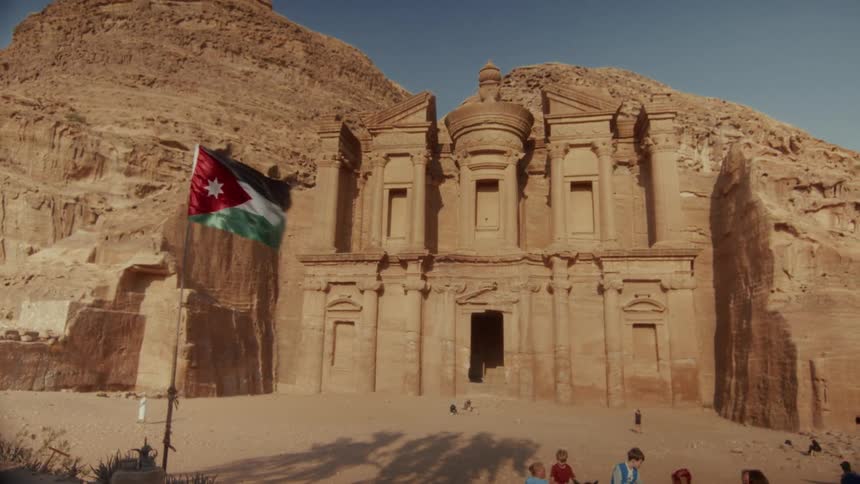 The magnificent Ad-Deir Monastery carved into the sandstone cliffs of Petra, Jordan, with the Jordanian flag waving in the foreground