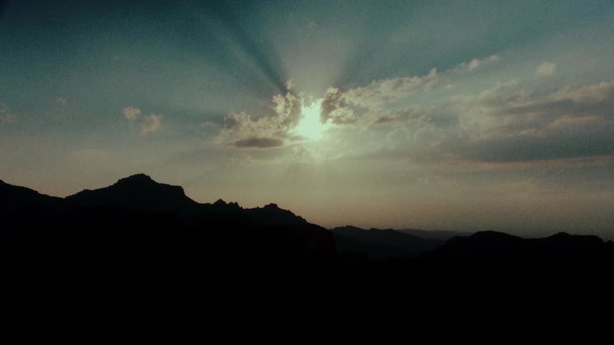 A dramatic timelapse captures brilliant sun rays spreading from behind clouds over a silhouetted mountain range at dusk