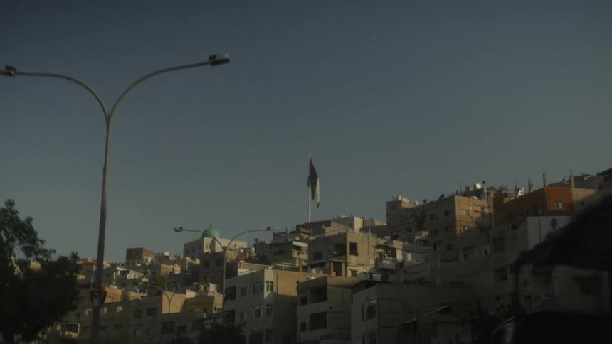 A slow-motion driving shot reveals the hillside cityscape of Amman, Jordan, with the national flag waving against a clear sky