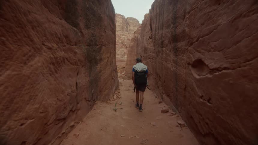 A tourist with a backpack walks in slow motion through a narrow and tall red rock canyon
