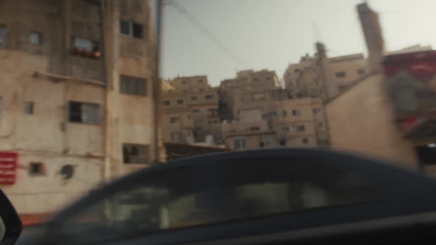 A point-of-view shot from a moving vehicle captures the weathered buildings and storefronts of a city in the Middle East