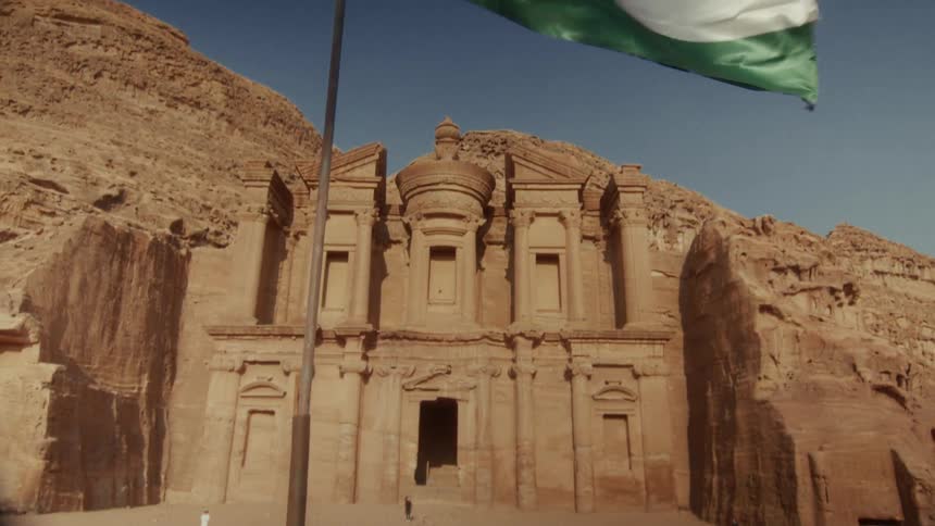 The majestic Jordanian flag waves in the wind as the camera tilts down to reveal the ancient Monastery in Petra